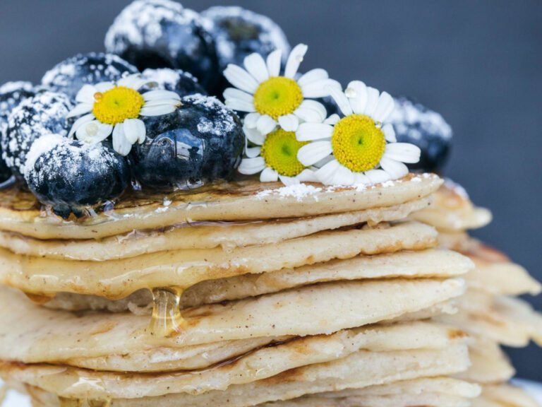 A stack of fluffy pancakes topped with fresh blueberries, honey, and white daisy flowers, perfect for a modern kitchen lifestyle presentation.
