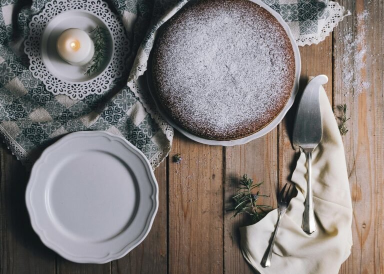 A round cake dusted with powdered sugar on a rustic wooden table, accompanied by a candle, plates, and cutlery for a cozy kitchen atmosphere.