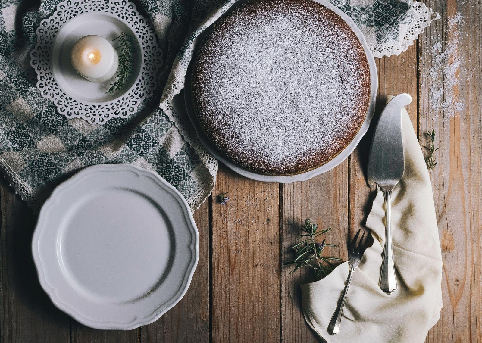 A round cake dusted with powdered sugar on a rustic wooden table, accompanied by a candle, plates, and cutlery for a cozy kitchen atmosphere.