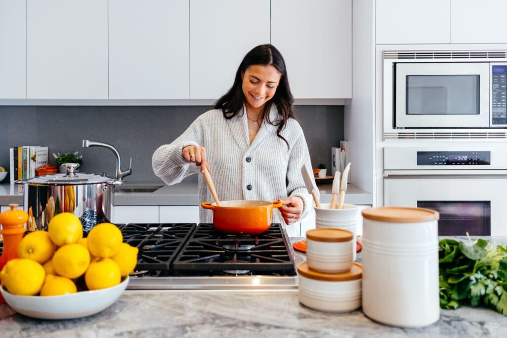 A woman cooking in a modern white Einbauküchen Tessin kitchen with high-end appliances, marble countertops, and organized storage solutions.