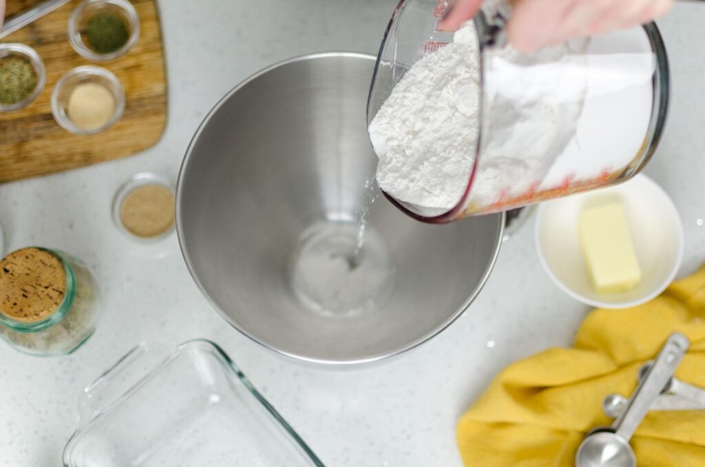 Top view of a hand pouring flour from a measuring cup into a stainless steel bowl on a modern kitchen countertop for Einbauküchen Tessin planning.