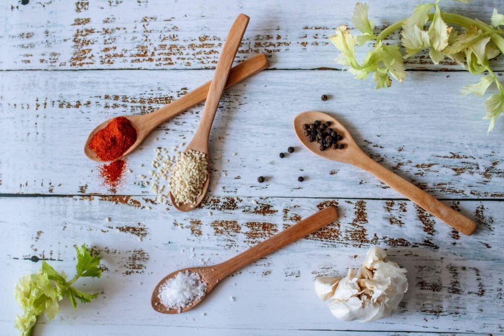 Four wooden spoons filled with spices like paprika, sesame seeds, peppercorns, and salt on a rustic white wooden background for Einbauküchen Tessin.